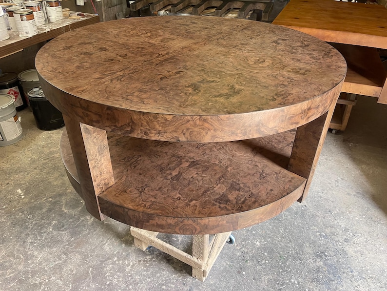 May include: A round, two-tiered table crafted from brown wood with a prominent burl grain pattern. The table features a circular top surface and a matching circular lower shelf, both supported by thick, vertical wooden legs in a matching finish.