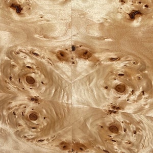 May include: A light beige wood veneer sheet featuring a symmetrical book-matched pattern. The surface displays a complex arrangement of dark brown circular burls and swirling grain lines, creating a natural, organic texture across the flat panel.
