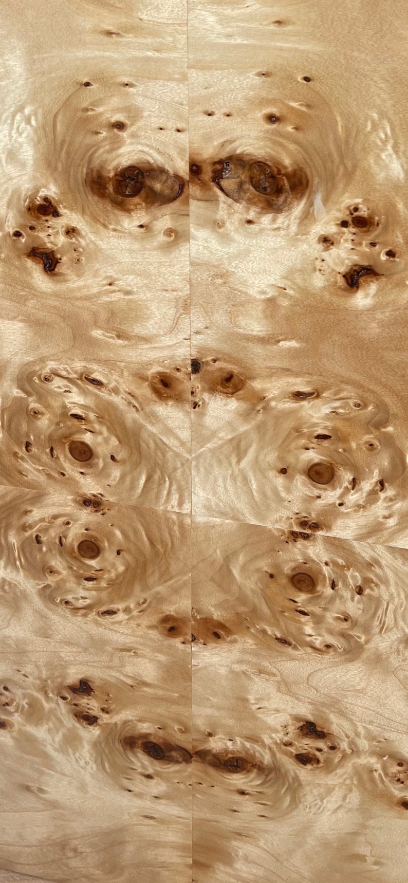 May include: A light beige wood veneer sheet featuring a symmetrical book-matched pattern. The surface displays a complex arrangement of dark brown circular burls and swirling grain lines, creating a natural, organic texture across the flat panel.