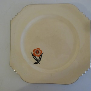 May include: A square, cream-colored ceramic plate with a scalloped edge. The plate features a stylized orange flower with black stems and leaves.