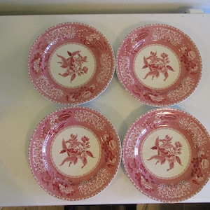 May include: Four matching pink and white decorative plates. Each plate features a central floral design surrounded by a detailed border. The plates have a vintage aesthetic, suitable for serving or display.