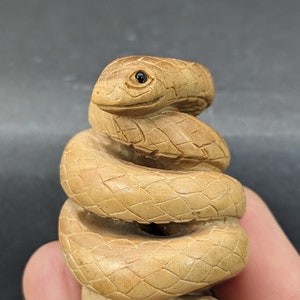 May include: A hand-carved wooden snake figurine, coiled in a spiral shape. The snake is a light brown color with a detailed, textured surface.