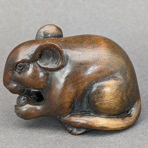 Peut inclure: Une petite sculpture en bois sculptée de manière complexe d'une souris, avec une finition brune. La souris est représentée en position assise, la queue enroulée autour de son corps. La souris a une apparence réaliste, avec des détails tels que ses yeux, ses oreilles et ses moustaches.