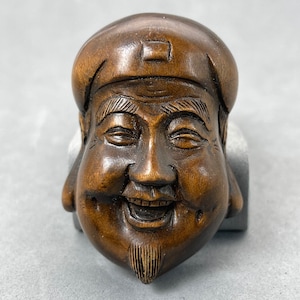 May include: A carved wooden figurine of a smiling, bearded man wearing a hat. The figurine is brown and has a detailed face.