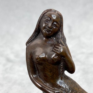 Peut inclure: Une sculpture en bois brun foncé d'une sirène avec une longue queue flottante et un visage souriant.