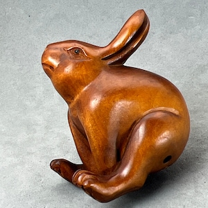 May include: A hand-carved wooden rabbit figurine with a reddish-brown finish. The rabbit is sitting with its ears perked up and its tail tucked behind its legs.