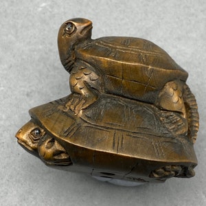 Op de afbeelding: Een gesneden houten sculptuur van twee schildpadden, een op de andere. De bovenste schildpad heeft zijn kop naar de zijkant gedraaid, terwijl de onderste schildpad zijn kop heeft ingetrokken. De sculptuur is donkerbruin van kleur en heeft een gladde, gepolijste afwerking.
