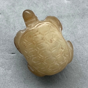 Puede incluir: Pequeña figura de tortuga de color claro con detalles grabados en el caparazón. La tortuga está en posición de gateo, con la cabeza y las extremidades visibles. La figura tiene un aspecto liso y pulido.