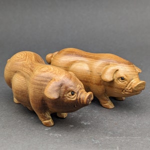 Könnte beinhalten: Zwei hölzerne Schweinefiguren, geschnitzt in einer hellbraunen Farbe mit detaillierten Merkmalen. Die Schweine sind nach vorne gerichtet, mit leicht angehobenen Schnauzen.