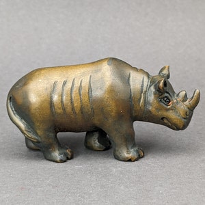 May include: A small, intricately carved wooden rhinoceros figurine. The figurine is painted in a dark brown color with a glossy finish. The rhinoceros is depicted in a walking pose with its head turned to the side.