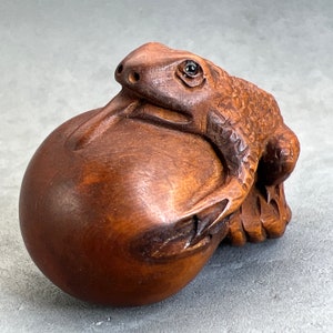 Peut inclure: Une sculpture en bois d'une grenouille tenant une boule ronde. La grenouille est sculptée en détail, avec une peau texturée et un grand œil. La boule est lisse et ronde.