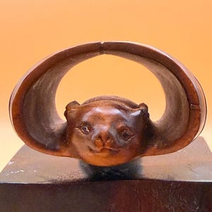 May include: A carved wooden netsuke sculpture depicting a bat with its wings wrapped around its head. The wood has a polished, dark brown finish with visible grain. The bat features detailed facial features, including eyes and ears, and is mounted on a dark, rectangular wooden base.