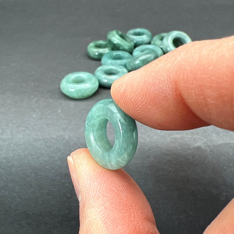 May include: A pile of light green jade donut beads. The beads are smooth and polished, with a slightly translucent appearance.
