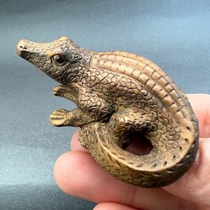 May include: A detailed wood carving of a crocodile with a textured, scaly surface. The crocodile is curled into a circle, with its tail tucked under its body.
