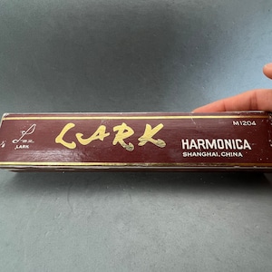 以下が含まれることがあります： 「LARK」という文字が金色で書かれた赤と金の箱。箱には「HARMONICA SHANGHAI, CHINA」という文字が黒で書かれており、モデル番号「M1204」も印刷されています。
