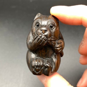 Puede incluir: Una talla de madera marrón oscuro de un mono que se tapa la boca con la mano. El mono está sentado con las piernas cruzadas y sosteniendo un objeto pequeño en su otra mano.