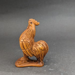 Peut inclure: Une sculpture en bois d'un coq debout sur une base. Le coq est brun et présente une sculpture détaillée des plumes. Le coq est tourné vers la droite.