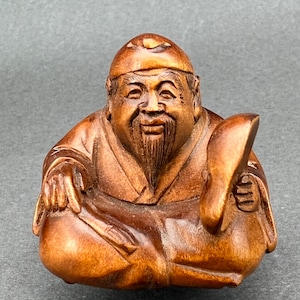 May include: A small wooden sculpture of a smiling, bearded man wearing a hat and traditional clothing. He is holding a fan in his left hand and a round object in his right hand.