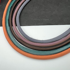 Puede incluir: Cinco cordones de tela de diferentes colores, dispuestos en un semicírculo. Los cordones son naranja, verde azulado, marrón, malva y gris.