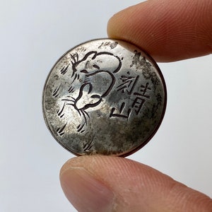 Könnte beinhalten: Eine silberne Münze mit einem Rattenmotiv und chinesischen Schriftzeichen. Die Münze ist leicht abgenutzt und hat eine Patina.