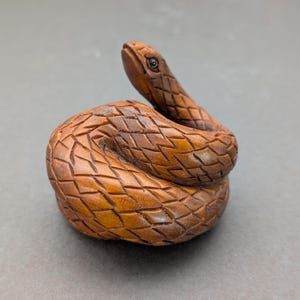 Peut inclure: Sculpture en bois détaillée d'un serpent enroulé. Le serpent est sculpté avec des écailles complexes et présente une tête relevée avec un œil détaillé. Le bois a une teinte brune chaude, et la sculpture est posée sur un fond neutre.