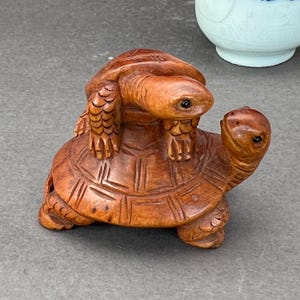 May include: A wooden sculpture of two turtles, one atop the other. The larger turtle has a detailed shell pattern and the smaller turtle is perched on its back. The sculpture is a warm brown color and has visible wood grain.