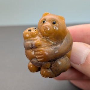 May include: A detailed, hand-carved figurine of a bear embracing a smaller animal. The bear and the smaller animal are a warm brown color with dark brown details. The figurine is small and appears to be made of wood.
