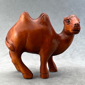 May include: A hand-carved wooden camel figurine with a reddish-brown color. The camel has two humps and is standing with its head turned to the side.