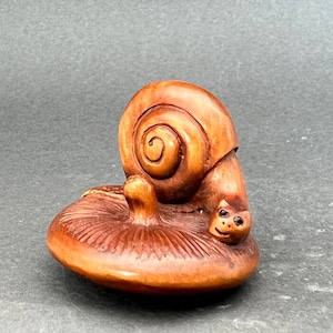 May include: A wooden sculpture of a snail with a smiling face, sitting on a mushroom. The snail is brown and has a detailed shell. The mushroom is also brown and has a textured surface.