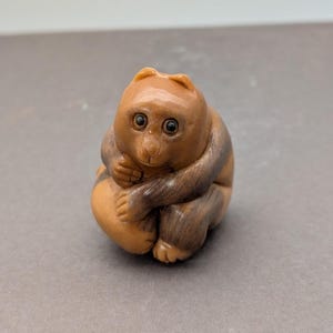 Puede incluir: Figura de madera tallada de un mono en posición sentada, abrazando sus rodillas. El mono tiene un cuerpo marrón claro con marcas marrones oscuras en los brazos y la espalda. Los ojos son negros, y el estilo es detallado.