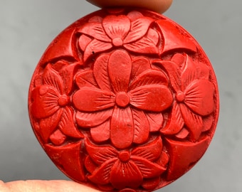 Può includere: Un ciondolo rotondo di colore rosso, intagliato con un motivo floreale. Il ciondolo è realizzato in un materiale liscio e lucido e presenta un motivo floreale dettagliato.
