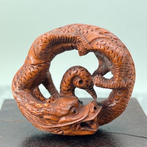Puede incluir: Una talla de madera detallada de un dragón enroscado en un círculo, con la cabeza y la cola uniéndose. El dragón está tallado intrincadamente con escamas y detalles, y sus ojos son prominentes.