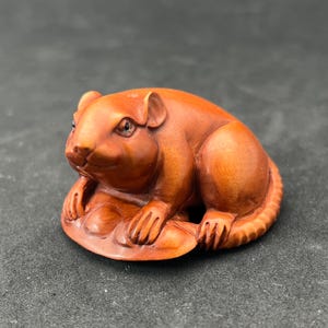 Puede incluir: Una figurita de rata de madera tallada a mano, sentada sobre una hoja. La rata es marrón y tiene ojos negros. La figurita es un netsuke japonés tradicional.
