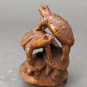 Op de afbeelding: Een gedetailleerde houten sculptuur van drie schildpadden die op elkaar zijn gestapeld. De schildpadden hebben een warme bruine kleur, met ingewikkelde schildpatronen en zichtbare gezichtskenmerken. De sculptuur staat tegen een neutrale grijze achtergrond.