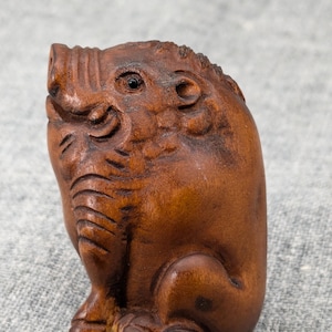 May include: A small, intricately carved wooden sculpture of a boar. The boar is brown and has a detailed face with a small black eye. The sculpture is sitting on a gray fabric surface.
