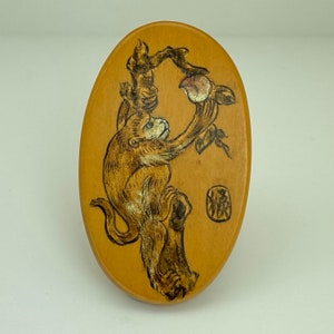 May include: A wooden oval ornament with a hand-painted design of a monkey reaching for a peach on a branch. The ornament is light brown with black and white details. The word "猿" is written in black ink on the ornament.