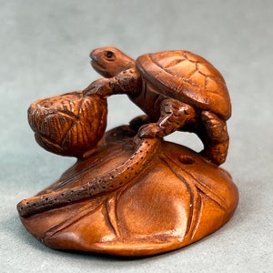 Op de afbeelding: Een handgesneden houten schildpadbeeldje dat op een lotusblad zit en een lotusknop in zijn voorpoten vasthoudt. De schildpad is bruin en heeft gedetailleerde snijwerk op zijn schild en poten. Het lotusblad is ook gesneden met ingewikkelde details.