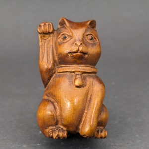 May include: A wooden Maneki-neko figurine, a traditional Japanese cat statue, with its right paw raised in a beckoning gesture. The cat is carved from light brown wood, with a collar and bell detail. The figurine is sitting upright.