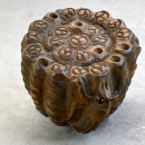 Puede incluir: Un objeto de madera marrón oscuro con un diseño tallado. El objeto tiene una parte superior circular con múltiples agujeros y un agujero central. La parte inferior del objeto está tallada con un diseño detallado.