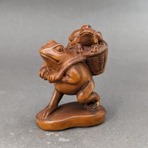 Puede incluir: Una talla de madera de una rana que lleva una cesta de ranas más pequeñas en su espalda. La rana es marrón y tiene una apariencia detallada y realista.