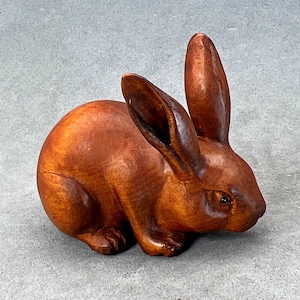 May include: A hand-carved wooden rabbit figurine in a brown color. The rabbit is sitting with its ears up and its eyes closed.