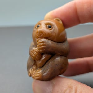 Puede incluir: Figurilla detallada, tallada a mano, de una criatura parecida a un oso. La escultura es principalmente marrón con acentos más claros, y la criatura se representa sosteniendo sus patas en su boca. La figurilla tiene un acabado liso y pulido.