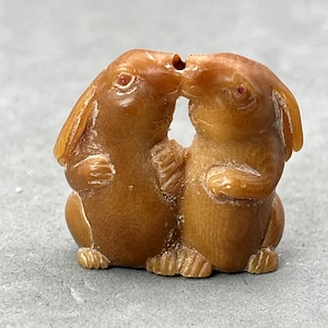 May include: A hand-carved wooden sculpture of two rabbits kissing. The rabbits are brown and have long ears. The sculpture is a traditional Japanese netsuke.
