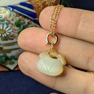 May include: A gold-colored necklace featuring a pendant carved from a light-colored stone. The pendant is shaped like a small pouch with intricate carved details. The necklace chain is a delicate, linked design.