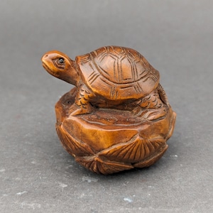 Op de afbeelding: Een handgesneden houten schildpadbeeldje dat op een lotusbloembasis staat. De schildpad is bruin en heeft gedetailleerde houtsnijwerk op zijn schild en poten. De lotusbloembasis is ook bruin en heeft ingewikkelde houtsnijwerk.