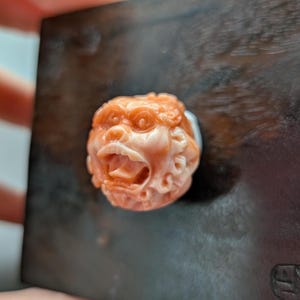 May include: A detailed carving of a mythical creature's head, featuring an open mouth and intricate facial details. The sculpture is a light orange color and appears to be made of a stone-like material. The carving is set against a dark background.