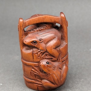 Peut inclure: Une sculpture en bois de deux grenouilles assises sur un seau. Les grenouilles sont sculptées dans un style réaliste et présentent des détails précis. Le seau est en bois et possède une poignée.