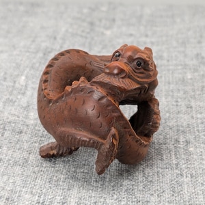 May include: A dark brown wooden carving of a dragon curled in on itself. The dragon's eyes are black and the carving is detailed with scales.
