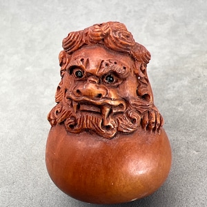 Peut inclure: Une sculpture en bois d'une créature mythique à l'expression féroce. La créature a un corps rond et un visage détaillé avec des dents acérées et de grands yeux. La sculpture est peinte dans des tons de brun et a une surface texturée.