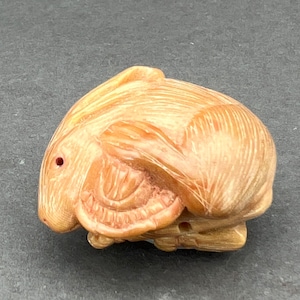 May include: A hand-carved wooden rabbit figurine with a light brown color and a detailed design. The rabbit is curled up in a sleeping position with its head resting on its paws.